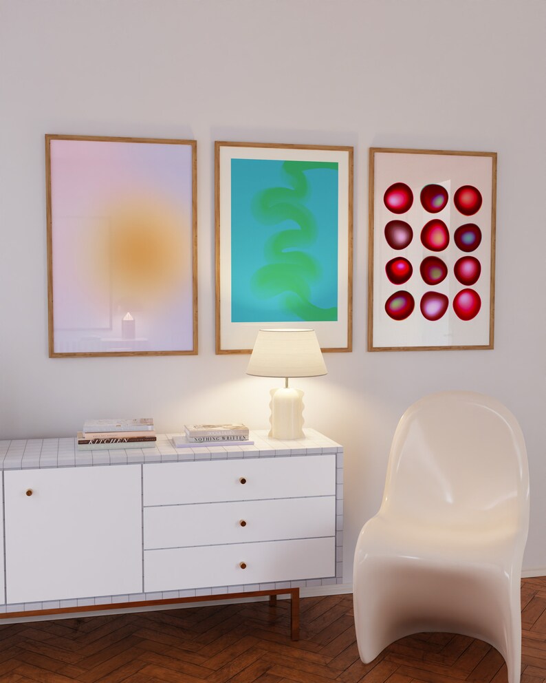 May include: Three framed abstract art prints on a white wall above a white dresser with three drawers. The prints are in a light pink, blue, and white color scheme. The dresser has a white top and gold knobs. A white lamp sits on the dresser. A white chair is in the foreground.