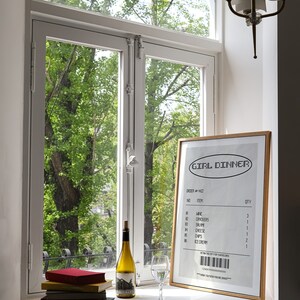 May include: A window-lit scene features a framed "GIRL DINNER" receipt, a wine bottle, a wine glass, and a stack of books. The receipt lists items like wine, crackers, and ice cream. The window overlooks lush green trees.