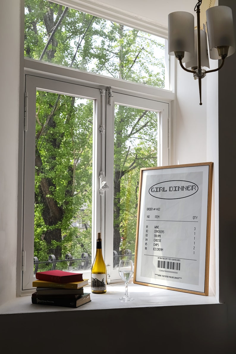 May include: A window-lit scene features a framed "GIRL DINNER" receipt, a wine bottle, a wine glass, and a stack of books. The receipt lists items like wine, crackers, and ice cream. The window overlooks lush green trees.
