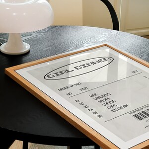 May include: A framed print with the text "GIRL DINNER" in a retro style. The print resembles a receipt, listing items like wine, crackers, salami, cheese, chips, and ice cream. The frame is made of light-colored wood, and the print is on a black table.