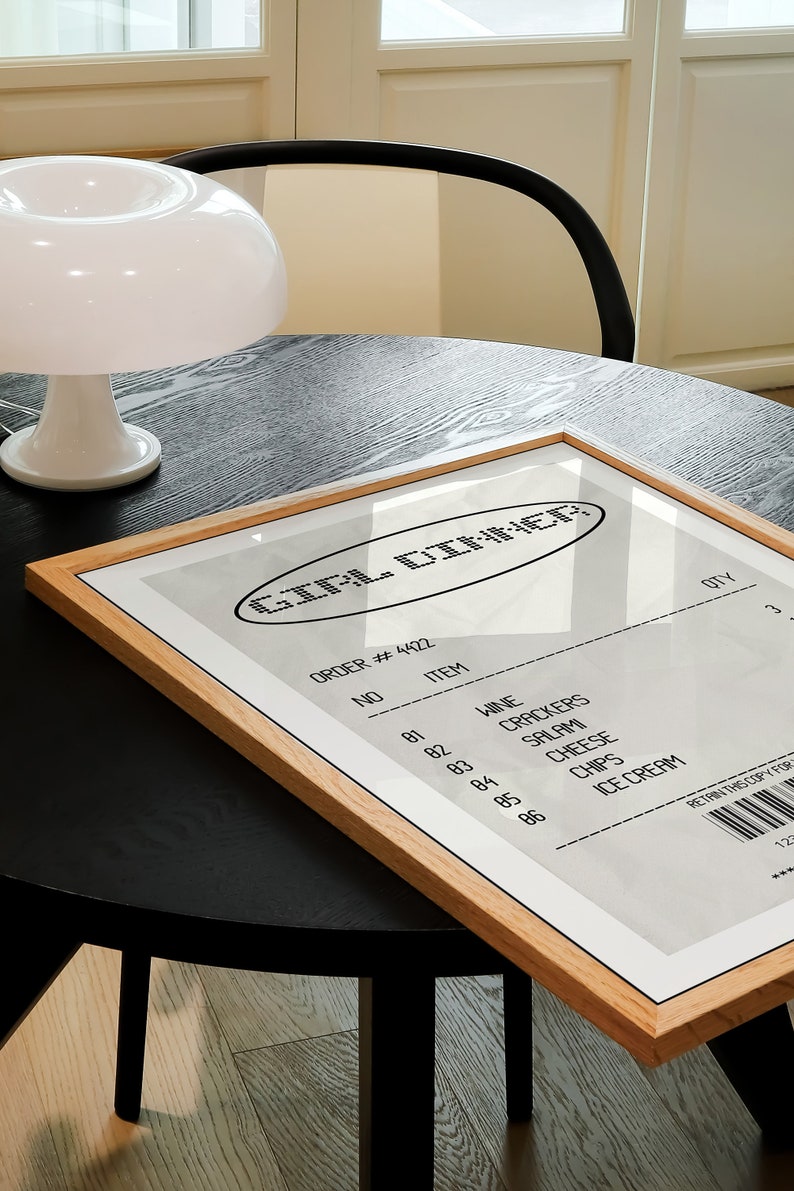 May include: A framed print with the text "GIRL DINNER" in a retro style. The print resembles a receipt, listing items like wine, crackers, salami, cheese, chips, and ice cream. The frame is made of light-colored wood, and the print is on a black table.