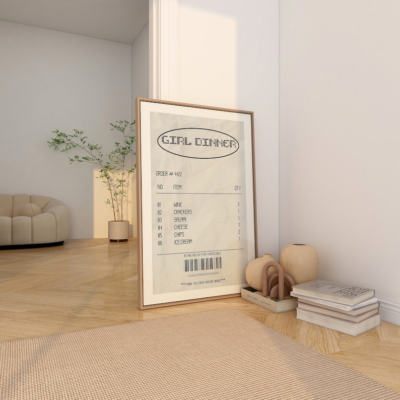 May include: A framed print with the words "GIRL DINNER" at the top. The print resembles a receipt, listing items like wine, crackers, salami, cheese, chips, and ice cream. The frame is a light brown color.