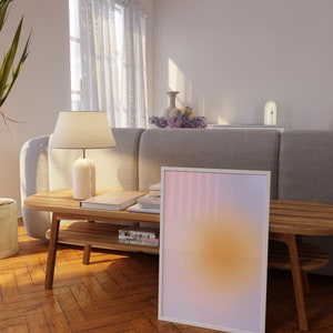 May include: A white framed print with a gradient of pink, purple, and yellow colors. The print is leaning against a wooden table with books and a lamp on top. The table is in a living room with a gray sofa and white curtains.