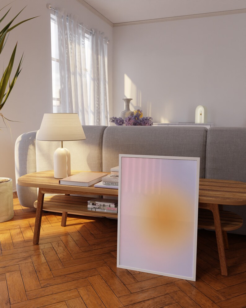 May include: A white framed print with a gradient of pink, purple, and yellow colors. The print is leaning against a wooden table with books and a lamp on top. The table is in a living room with a gray sofa and white curtains.