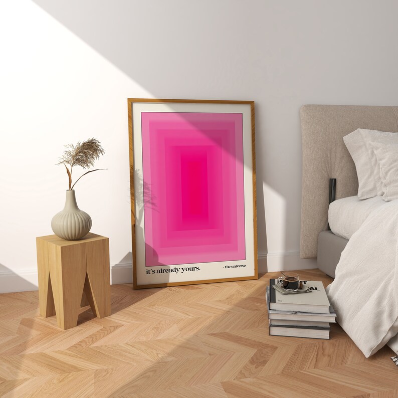 May include: A framed pink and white abstract geometric print with the text "it's already yours - the universe" at the bottom. The print is hanging on a white wall above a wooden stool.