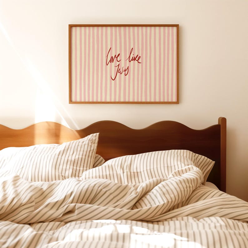 May include: A framed artwork with a pink and cream striped background and the handwritten text "Love like Jesus" in red. The artwork hangs above a bed with striped bedding and a wooden headboard.