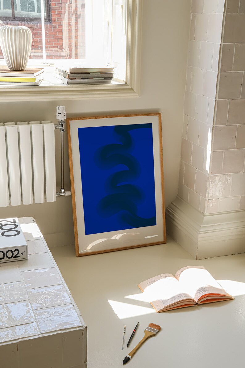 May include: A framed abstract print with a blue background and a dark blue, swirling, abstract design. The print is hanging on a white wall next to a white radiator.