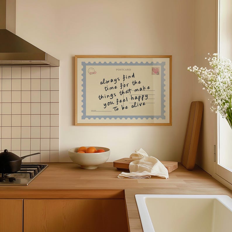 May include: A framed print with the text "always find time for the things that make you feel happy To be alive." The print is designed to look like a postcard with a light blue border and a postage stamp. The frame is made of light wood.