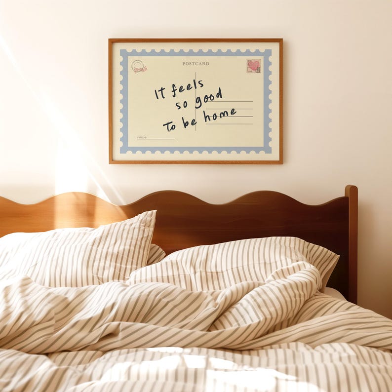 May include: A framed postcard print with the text "It feels so good to be home" hangs above a bed. The postcard has a blue border and a stamp design. The bed has striped bedding and a wooden headboard.