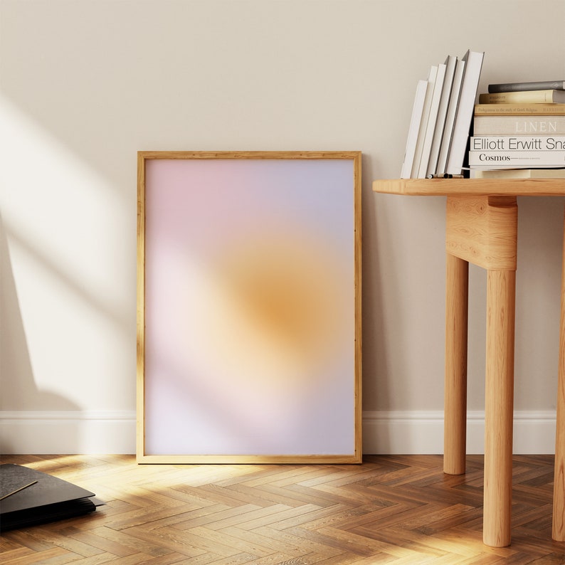 May include: A framed abstract print with a gradient of pink, orange, and yellow colors. The print is hanging on a white wall above a wooden table with a stack of books.