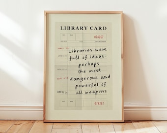 Puede incluir: Impresión enmarcada con un diseño de tarjeta de biblioteca vintage. El texto dice: "Las bibliotecas estaban llenas de ideas, quizás el arma más peligrosa y poderosa de todas". La impresión tiene un fondo beige y un marco de madera.
