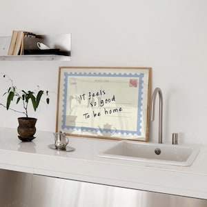 May include: A framed postcard print with the text "It feels so good to be home" in a kitchen setting. The print has a blue border and a postage stamp design. A small potted plant and a silver teapot are also visible.