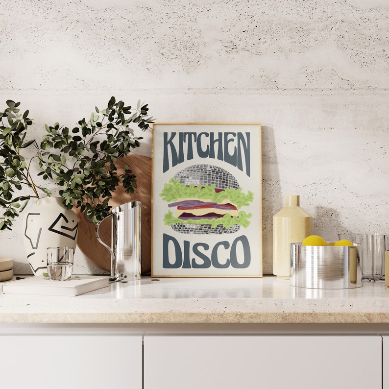 Pu&ograve; includere: Una stampa incorniciata con le parole "KITCHEN DISCO" sopra e sotto un'illustrazione di hamburger a palla da discoteca. L'hamburger ha lattuga, una fetta di cipolla rossa e formaggio. La stampa &egrave; esposta su un piano di lavoro bianco.