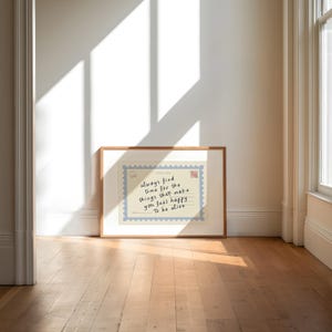 May include: A framed print with a light wood frame rests against a white wall. The print features a postcard design with the handwritten text: "always find time for the things that make you feel happy. To be alive."