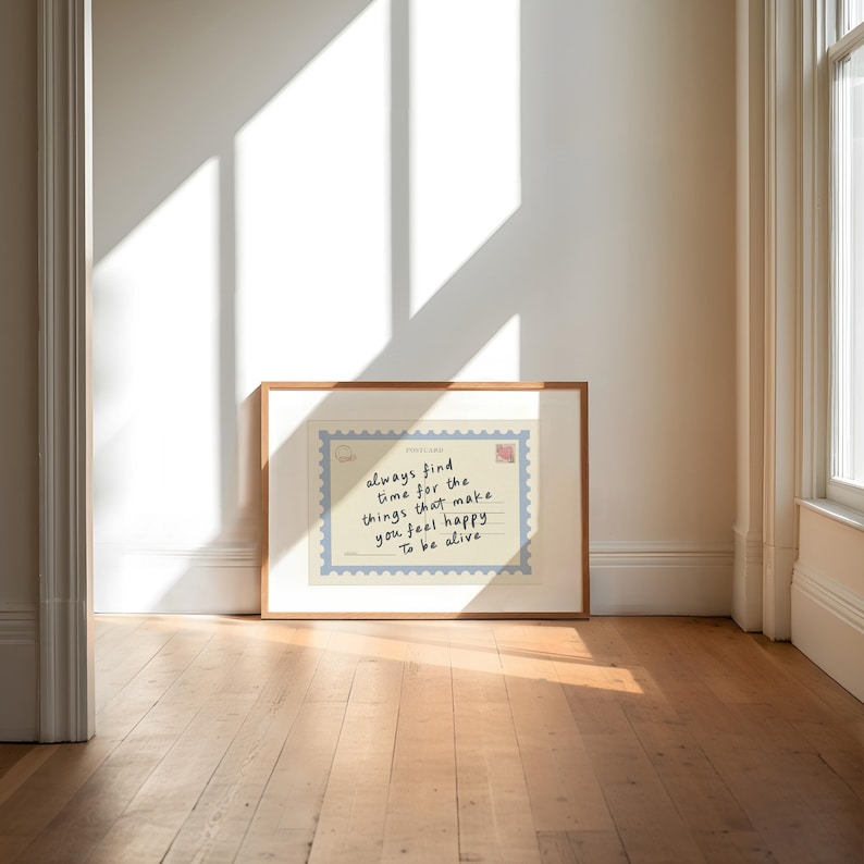 May include: A framed print with a light wood frame rests against a white wall. The print features a postcard design with the handwritten text: "always find time for the things that make you feel happy. To be alive."