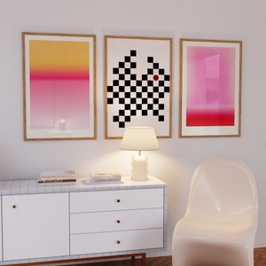 May include: Three framed abstract art prints with a white dresser and a white chair in the background. The prints are a gradient of pink and yellow, a black and white chequered pattern, and a gradient of pink and red.
