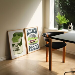 Pu&ograve; includere: Due stampe artistiche incorniciate con cornici in legno. Una dice "EAT YOUR GREENS" con una grafica di verdure. L'altra dice "KITCHEN DISCO" con una grafica di hamburger a palla da discoteca. Una sedia e un tavolo neri sono in primo piano.