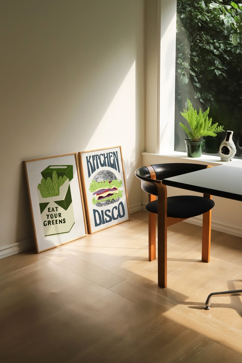 Pu&ograve; includere: Due stampe artistiche incorniciate con cornici in legno. Una dice "EAT YOUR GREENS" con una grafica di verdure. L'altra dice "KITCHEN DISCO" con una grafica di hamburger a palla da discoteca. Una sedia e un tavolo neri sono in primo piano.