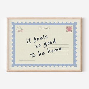 May include: A framed postcard print with a light beige background and a blue border. The postcard reads "It feels so good to be home" in handwritten black script. A small heart-shaped stamp is in the upper right corner.