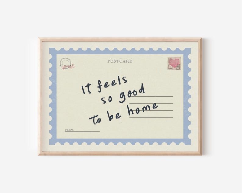 May include: A framed postcard print with a light beige background and a blue border. The postcard reads "It feels so good to be home" in handwritten black script. A small heart-shaped stamp is in the upper right corner.