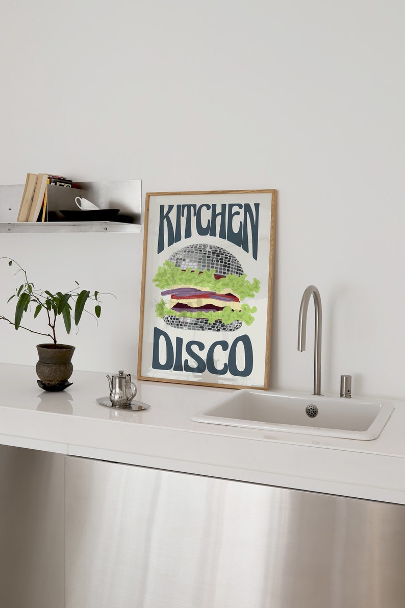 Pu&ograve; includere: Una stampa artistica incorniciata con un design a forma di hamburger. L'hamburger &egrave; composto da una palla da discoteca, lattuga, pomodoro e cipolla. Le parole "KITCHEN DISCO" sono scritte in blu scuro. La stampa ha una cornice in legno.