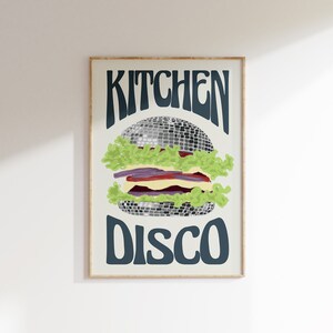 Pu&ograve; includere: Una stampa artistica incorniciata con un design di hamburger stravagante. L'hamburger &egrave; raffigurato con una palla da discoteca come polpetta, circondata da lattuga, pomodoro e altri condimenti. Le parole "KITCHEN DISCO" sono scritte sopra e sotto l'hamburger.