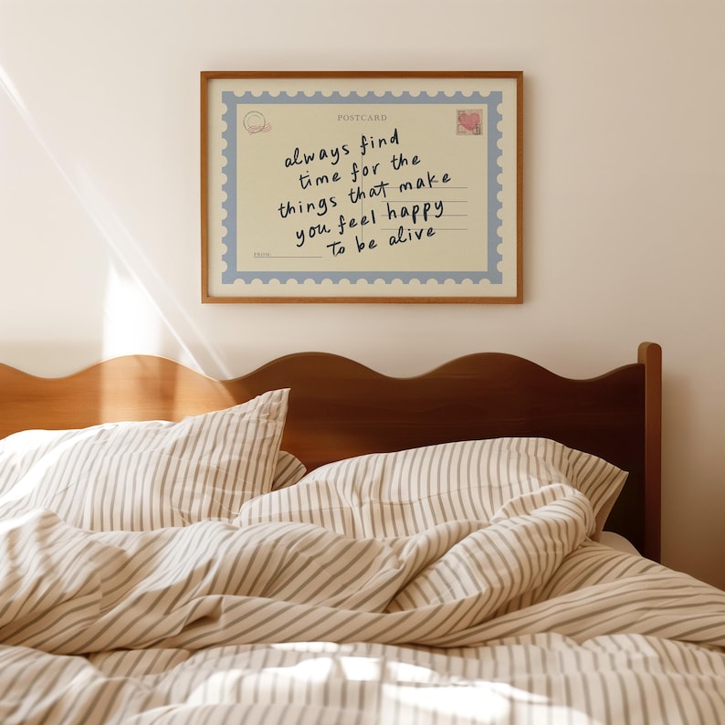 May include: A framed postcard with the text "always find time for the things that make you feel happy To be alive." The postcard is light beige with a blue border and a wooden frame. The postcard is above a bed with striped bedding.