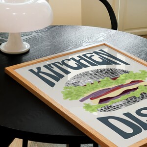 Pu&ograve; includere: Una stampa incorniciata con un'illustrazione di hamburger e le parole "KITCHEN DISH" in grassetto. L'hamburger presenta lattuga, pomodoro e un patty grigio. La cornice &egrave; in legno chiaro e la stampa &egrave; su un tavolo.