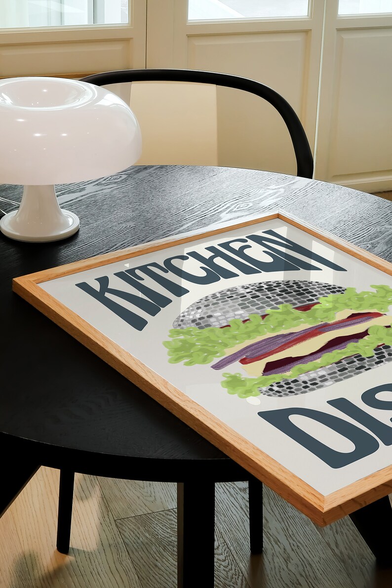 Pu&ograve; includere: Una stampa incorniciata con un'illustrazione di hamburger e le parole "KITCHEN DISH" in grassetto. L'hamburger presenta lattuga, pomodoro e un patty grigio. La cornice &egrave; in legno chiaro e la stampa &egrave; su un tavolo.