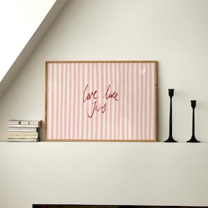 May include: A framed print with a pink and white striped background and the words "love like Jesus" in red script. Two black candle holders stand to the right of the print. A stack of books sits to the left.