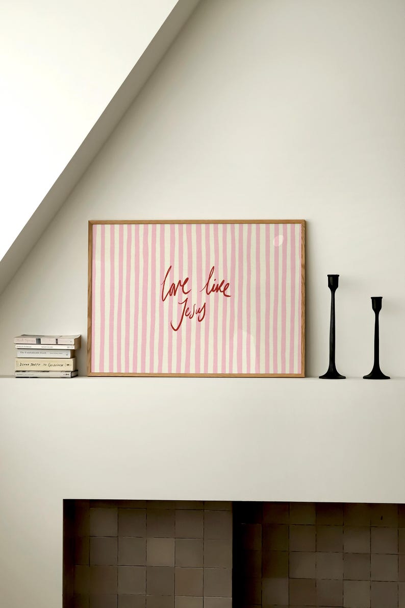 May include: A framed print with a pink and white striped background and the words "love like Jesus" in red script. Two black candle holders stand to the right of the print. A stack of books sits to the left.