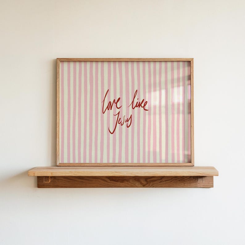 May include: A framed print with a pink and white striped background and the handwritten text "love like Jesus" in red. The frame is a light brown color and sits on a wooden shelf.