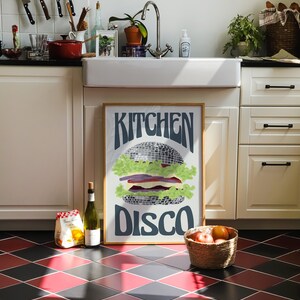 Pu&ograve; includere: Un poster incorniciato con le parole "Kitchen Disco" e un'illustrazione di hamburger a palla da discoteca. L'ambiente della cucina comprende un lavandino bianco, una bottiglia di vino e un cesto di frutta. Il pavimento ha un motivo a rombi rossi e neri.