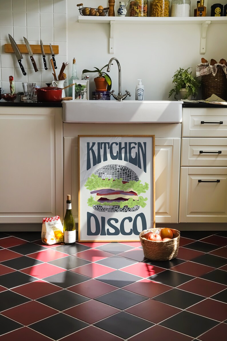 Pu&ograve; includere: Un poster incorniciato con le parole "Kitchen Disco" e un'illustrazione di hamburger a palla da discoteca. L'ambiente della cucina comprende un lavandino bianco, una bottiglia di vino e un cesto di frutta. Il pavimento ha un motivo a rombi rossi e neri.