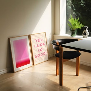 K&ouml;nnte beinhalten: Zwei gerahmte Drucke auf einem hellen Holzboden. Der Druck links ist ein Farbverlauf von Rosa und Rot. Der Druck rechts ist ein Gittermuster mit dem Text "YOU LOOK SO GOOD!" in Rosa.