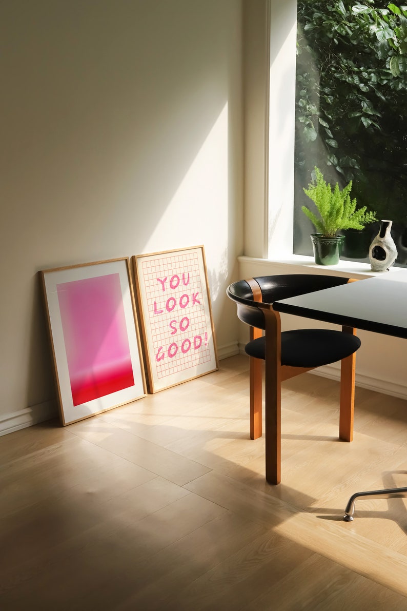 K&ouml;nnte beinhalten: Zwei gerahmte Drucke auf einem hellen Holzboden. Der Druck links ist ein Farbverlauf von Rosa und Rot. Der Druck rechts ist ein Gittermuster mit dem Text "YOU LOOK SO GOOD!" in Rosa.