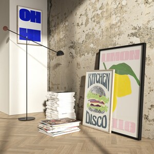 Pu&ograve; includere: Scena d'interni con stampe artistiche incorniciate con grafica e testo audaci, tra cui una stampa blu e bianca "OH HI", una stampa "Kitchen Disco" con hamburger e una stampa "Lemon". Una lampada da terra nera e pile di riviste completano la scena.