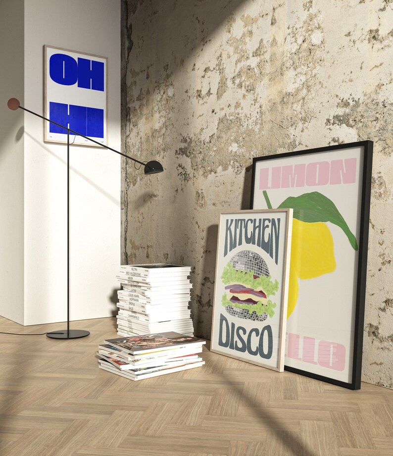 Pu&ograve; includere: Scena d'interni con stampe artistiche incorniciate con grafica e testo audaci, tra cui una stampa blu e bianca "OH HI", una stampa "Kitchen Disco" con hamburger e una stampa "Lemon". Una lampada da terra nera e pile di riviste completano la scena.