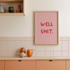 May include: A pink framed print with the text "WELL SHIT." in red lettering, hanging on a white tiled wall above a peach-coloured kitchen counter.
