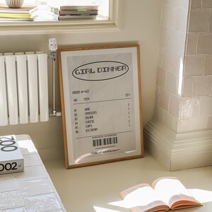 May include: A framed print with the words "GIRL DINNER" in a retro font, resembling a receipt. The print lists items like wine, crackers, and ice cream. The frame is wood-toned, and the print is propped against a wall.