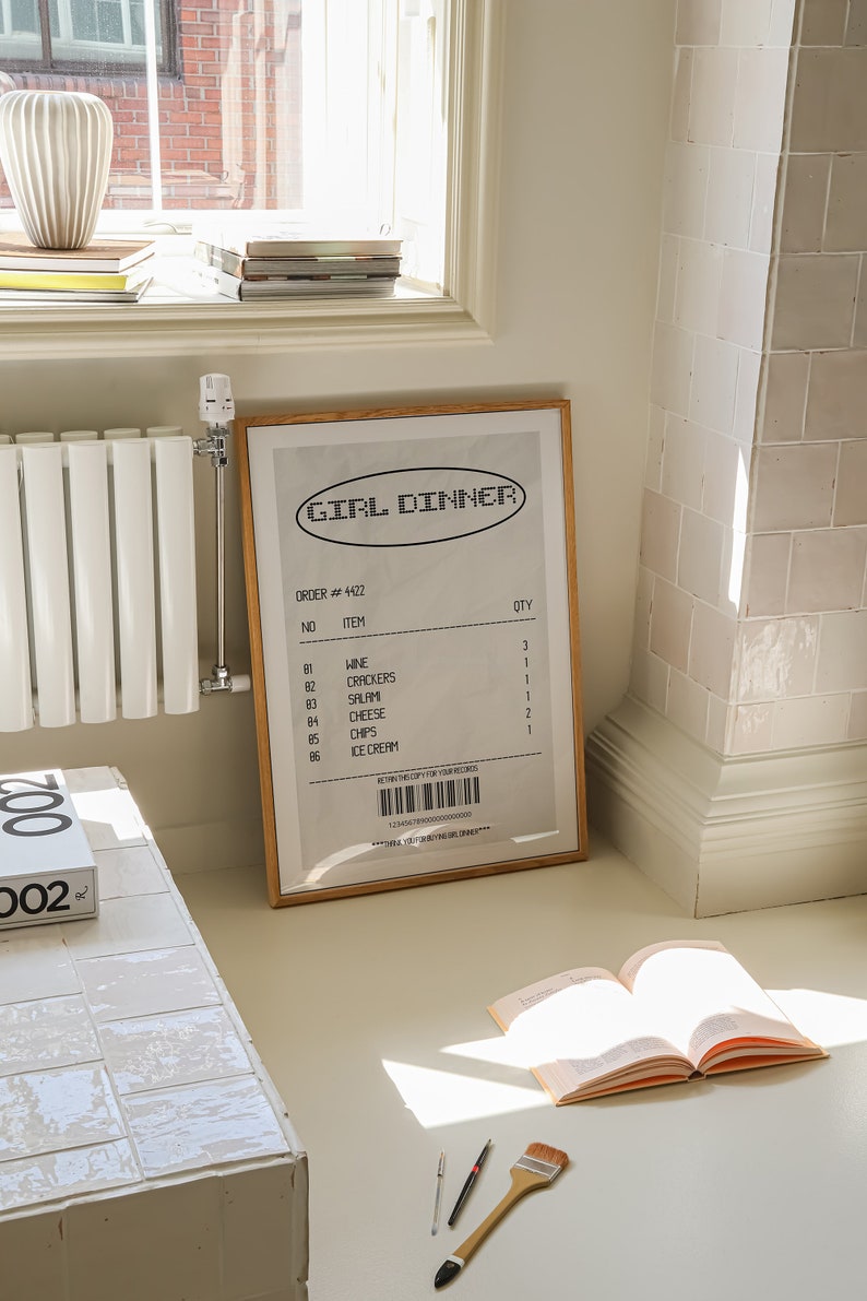 May include: A framed print with the words "GIRL DINNER" in a retro font, resembling a receipt. The print lists items like wine, crackers, and ice cream. The frame is wood-toned, and the print is propped against a wall.