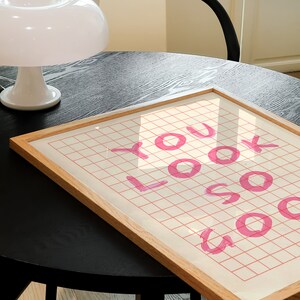 K&ouml;nnte beinhalten: Ein gerahmter Druck mit einem rosa Gittermuster und dem Text "YOU LOOK SO GOOD" in rosa.