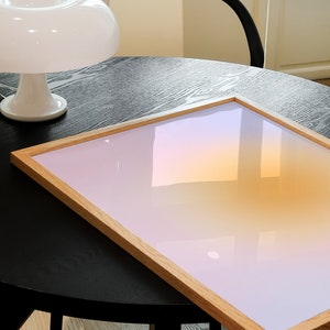 May include: A wooden framed picture with a gradient background of pink, orange, and yellow. The picture is sitting on a black table with a white lamp in the background.