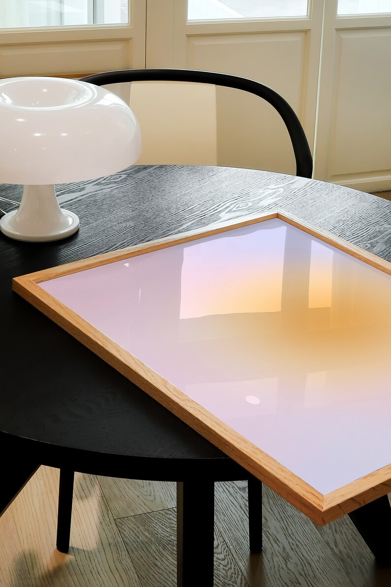May include: A wooden framed picture with a gradient background of pink, orange, and yellow. The picture is sitting on a black table with a white lamp in the background.