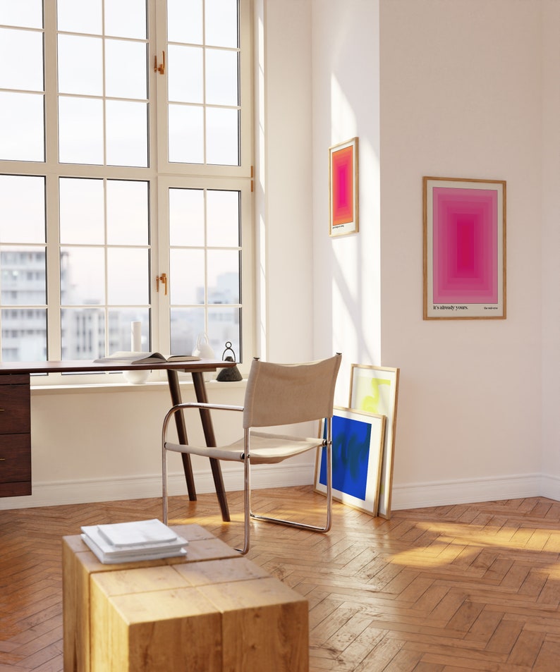 May include: A wooden desk with a chair and a stack of papers on a wooden stool. The desk is next to a large window with a view of a city. There are three framed prints on the wall, one with the text "It's already yours."