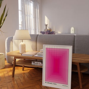 May include: A framed pink and white abstract art print with the text "It's already yours. - the universe" on a wooden table in a living room.