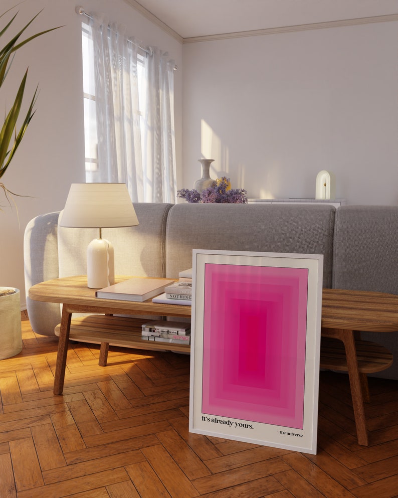 May include: A framed pink and white abstract art print with the text "It's already yours. - the universe" on a wooden table in a living room.