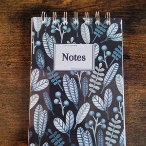 Puede incluir: Una pequeña libreta negra de espiral con un patrón de hojas azules y blancas y la palabra "Notes" en un recuadro blanco.