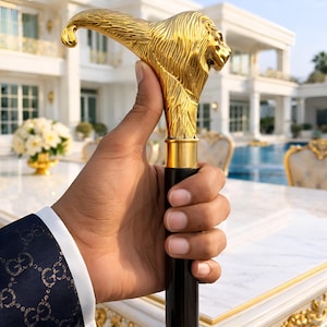 Op de afbeelding: Een gouden leeuwenkop wandelstok met een zwarte houten schacht. De manen van de leeuw zijn ingewikkeld gedetailleerd. De wandelstok wordt in een hand gehouden, met een marineblauw colbert met een gouden patroon op de achtergrond.