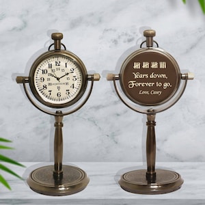 May include: A pair of antique brass table clocks with a round face. One clock has a traditional clock face with Roman numerals and the other clock has a personalized message that reads "Years down, Forever to go. Love, Casey".